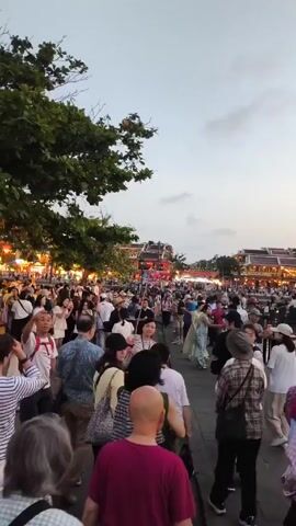 The video shows a large crowd of people walking down a street. There are buildings on either side of the street, and the scene is lit by streetlights and colorful lanterns. There are trees lining the street. The crowd consists of people of various ages, dressed in different styles of clothing. Some people are walking in groups, while others are walking alone. There is a general sense of activity and movement in the scene.