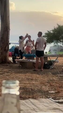 Here's a description of the video:

The scene is a campsite, likely near a body of water (possibly a lake or sea), as suggested by the glimpses of water in the background. Several people are visible around a picnic table. One woman is pointing, possibly directing or indicating something. Another woman is holding a bottle or can. A man stands with his hands on his hips, seemingly engaged in the conversation. Other people are moving around in the background, tending to camping equipment, including tents. The ground is covered in dry leaves and there's a tree to the left of the frame. In the foreground, slightly out of focus, is a glass bottle. The overall atmosphere appears relaxed, with the setting sun suggesting a pleasant evening.