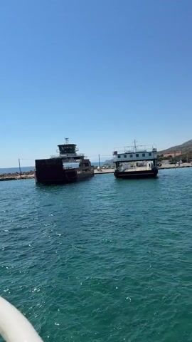 A ferryboat is departing from a dock. The ferryboat is dark brown and has a dark brown superstructure on the back. There are two decks on the superstructure, with windows on both. The front of the ferryboat is open, and there are vehicles visible on the deck. The ferryboat is moving away from the dock, and there is another ferryboat docked to the left of it. The second ferryboat is white and has a similar superstructure on the back. The water is blue and choppy, and the sky is clear and blue.
