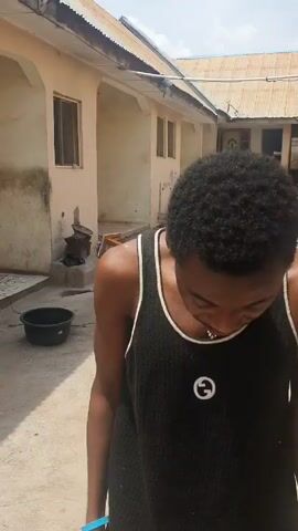 A young man wearing a black tank top with a white trim and a gold necklace stands in an outdoor space with buildings in the background. He's looking directly at the camera and speaking, gesturing with his hands. The area appears to be a courtyard or a similar open space, and the buildings seem to be of a simple, possibly residential style, with corrugated metal roofs. He has a bracelet on each wrist.