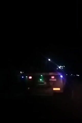 At night, the back of a semi-trailer truck is visible on a road. The truck is outfitted with red, green, and blue lights. Streetlights appear in the background, and the vehicle is in motion.