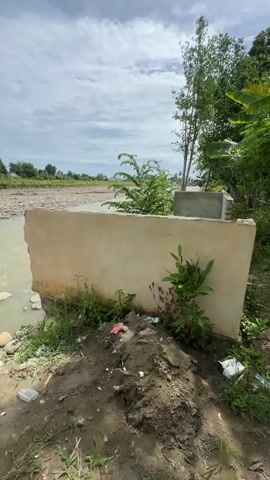 A video shows a partially constructed concrete structure beside a river. On the left, a river flows with a rocky bank. In the foreground, a large, flat concrete slab is partially submerged in the river and extends onto the bank. It is rough and unfinished, with some plants growing around its base. To the right of this slab, more unfinished concrete construction is visible, including brickwork and concrete walls. There are also scattered pieces of debris, including plastic bags, on the ground. In the distance, across the river, a flat, dry landscape is visible. Several trees are present on the right side of the frame, with lush green foliage. The sky is cloudy with patches of blue. At the 00:04 mark, three people are visible near the river bank, sitting or standing by the water.