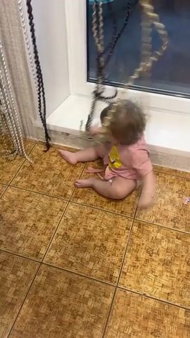 A baby is sitting on a tiled floor next to a window. The baby is wearing a pink shirt and a diaper. There are several strings of beads hanging from the window sill. The baby is reaching for and playing with the beads. The floor is made of square, brown tiles.
