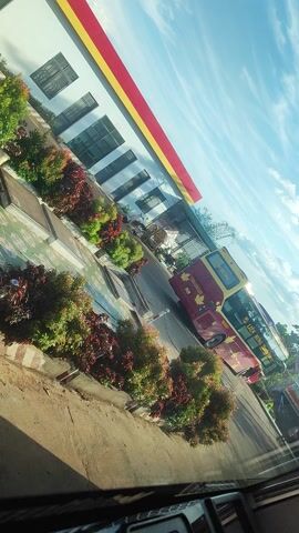 The media file shows a red and yellow bus parked in front of a white building with a red and yellow roof. The building appears to be a service station or a bus depot. There are rows of green and red bushes planted in front of the building. The sky is blue with some white clouds. The image is tilted, suggesting it was taken from a moving vehicle. The overall scene appears to be from a sunny day.

There are no people clearly visible in the image. The main objects are the bus, the building, and the landscaping. The scene depicts a bus, likely in transit or at a stop, at a roadside facility in Chư Sê, Vietnam. The time of day appears to be daytime, given the bright sunlight casting shadows. The weather seems pleasant, with a clear blue sky.

A notable detail is the text on the bus, which reads "Tien Hung" and appears to have Vietnamese characters. The bus itself is red and yellow, with a maple leaf logo. The building has a modern design with large windows. The landscaping consists of neatly trimmed bushes, creating a decorative border. The tilted angle of the camera adds a dynamic feel to the image, possibly indicating the perspective of a passenger on another vehicle. There are no strong emotional cues conveyed by the image; it is a straightforward depiction of a transportation hub.