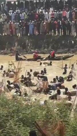 The video shows a large group of people standing on a bank overlooking a muddy body of water, while numerous individuals are wading through the water, using large nets to fish. There are also people in boats in the water.  The environment appears to be outdoors.