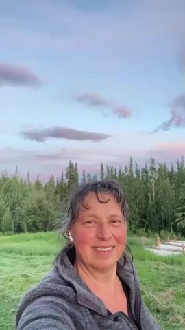 A woman stands in front of a treeline at dusk, smiling at the camera. She has grey hair pulled back and is wearing a grey hooded jacket. The sky above her is a gradient of blue, pink, and purple, with scattered clouds. The background consists of a field of green grass, with trees and some wooden structures visible behind her.