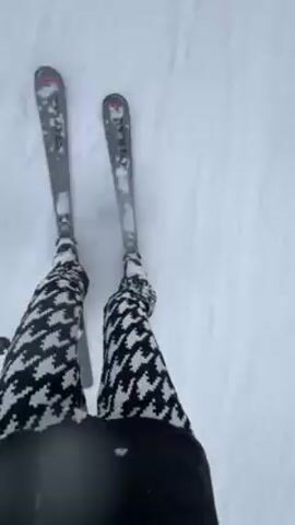 A woman skis downhill. She wears a black ski helmet with dark goggles, a black ski jacket with pink straps, and a blue neck warmer. She has dark hair braided down her back. She smiles at the camera. Her pants are patterned white and black. Her skis are gray and white.  The environment is snowy, with trees and mountains visible in the distance. Other skiers can be seen on the slope.  The weather is overcast and it is snowing lightly.