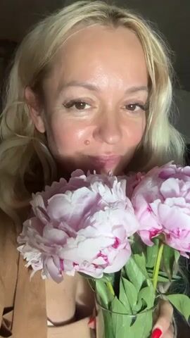 A close-up shot shows a woman with blonde hair and fair skin holding a bouquet of pink peonies. She is leaning in to smell the flowers, with her eyes looking towards the camera. The background is blurred, but appears to be indoors with some greenery visible. The woman is wearing a light brown jacket over a white top. Her makeup is visible, including eyeliner and blush. She has a mole on her cheek. Her fingernails are painted red. The video is silent.