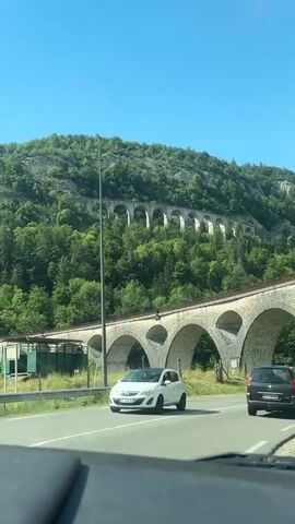 The video captures a view from inside a vehicle driving on a road that passes beneath a large stone arched viaduct. The viaduct is a prominent structure composed of numerous arches and is set against a backdrop of a lush, green, tree-covered hillside under a clear blue sky with scattered clouds. Sunlight illuminates the scene. In the foreground, the road is visible with white lane markings. A white car is seen on the left side of the road, and a black car is visible further ahead on the right side of the road, also driving away from the camera. A tall street light stands on the left side of the road, near the viaduct. The viaduct itself is constructed of stone blocks and features a railing along its top. In the distance, further up the hillside, another section of the viaduct or a similar structure with multiple arches can be seen winding along the slope. The overall impression is one of scenic natural beauty combined with impressive historical architecture.