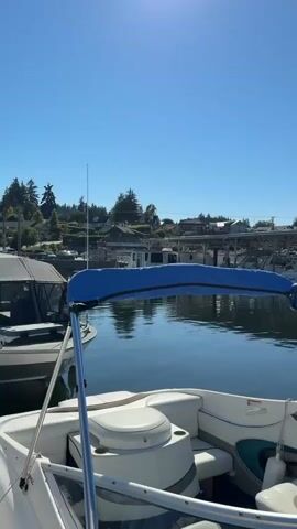 This video shows a view from a boat in a marina. The boat is white with blue accents and has a blue canopy. In the background, there is a row of covered boat slips with various boats docked inside. The water is calm and reflects the blue sky and the boats. There are some buildings and trees visible on the shore in the distance. The sun is shining, and the sky is clear.