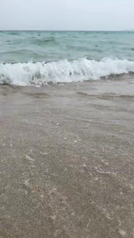 A low-angle, handheld shot shows the edge of a sandy beach where a gentle wave is rolling in. The water is a greenish-blue color with white foam on the crest of the wave. The sand is light brown and appears wet near the water's edge. The sky is overcast and appears a light gray. The video is composed of short, static clips.