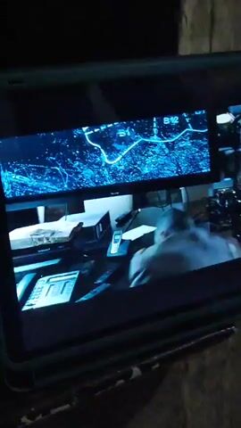 A man in a white shirt is seen from behind, looking at a scene on a screen that shows a man in a black suit and several other people standing in a room with large windows and pillars. The scene then cuts to a man in a dark room with multiple computer monitors, one of which displays a map with a timer counting down from 00:02. Another man, older with white hair, sits at a desk. The man in the dark room is speaking animatedly with his hands raised. The video is viewed on a tablet device.