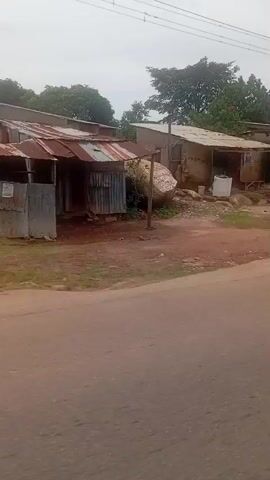 The video begins with a roadside scene in what appears to be a rural or semi-urban area. In the foreground, there is a dusty, reddish-brown dirt road. On the left side of the road, there is a small, makeshift structure with a corrugated metal roof, possibly a shop or stall. In front of this structure, several large appliances, including washing machines and refrigerators, are displayed for sale. Beyond the shop, there are trees and more structures in the background.

The camera then pans to the right, revealing a more established building with a fence and vegetation. This building appears to be a residential house with a covered porch area. More trees and foliage surround the property.

The camera continues to move, showing another roadside scene with more rudimentary structures, some appearing to be small shops or food stalls with tables and chairs set up outside. Trees are abundant in this area.

A silver sedan drives past the camera on the road.

Finally, the video shows a person in a blue uniform walking away from a small shack-like structure under a tree. The person is walking on a paved road in front of the shack. The shack appears to have some materials, possibly wood, piled near it. Red objects, possibly clothing or bags, are visible near the base of one of the trees on the left.