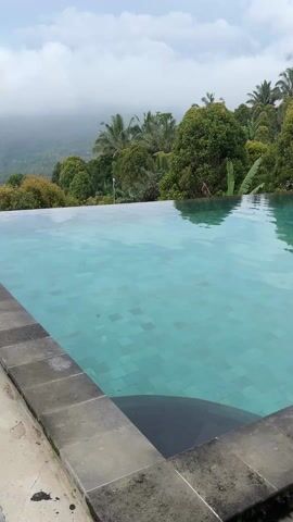 The video shows a swimming pool with an infinity edge. The pool is tiled in light blue or green squares. The edge of the pool is made of dark gray stone. Beyond the pool is a landscape of trees and mountains under a cloudy sky. There are lounge chairs and an umbrella next to the pool on the right side of the frame. The water is calm, reflecting the sky and trees.