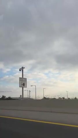 A white SUV is traveling on a road, with a concrete barrier on the right. Streetlights and palm trees are visible beyond the barrier. A modern building with a grey facade and windows is also visible in the distance. In the final frames, an Israeli flag is seen waving in front of the building. The sky is cloudy with patches of blue.