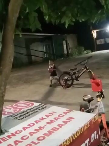 A young child is playing with a ball on the ground. The child is wearing a dark shirt and shorts. A bicycle and a small red toy cart are parked nearby. A tree is in the foreground, and a building with lights on is in the background. A banner with text in Indonesian is spread out on a table in front of the child. The scene appears to be outdoors at night.