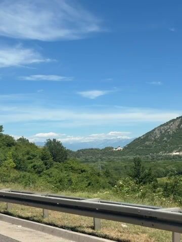 The video shows a scenic view from a moving vehicle. A metal guardrail is visible in the foreground, with lush green foliage and trees filling the middle ground. In the background, a mountain range is visible under a clear blue sky with scattered clouds. The sun appears to be shining, casting light on the landscape. No people or specific actions are captured in the video, beyond the implied movement of the camera.