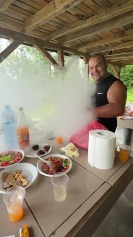 A man in a black tank top stands in an outdoor gazebo or shelter with a wooden beam ceiling, facing away from the camera. He is exhaling a large cloud of white smoke from what appears to be a vape or electronic cigarette. He has his arms outstretched and thumbs up as the smoke dissipates, revealing a table in the foreground laden with food and drinks. The table holds several plates of food, including salads, crackers, and what looks like meatballs or falafel. Drinks include water, orange soda, and clear beverages in plastic cups. A roll of toilet paper and a pink plastic bag are also on the table. The background shows trees and greenery, suggesting a natural outdoor setting.