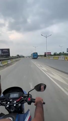 A first-person view of a person riding a blue Honda motorcycle on a multi-lane highway. The rider's hand is visible on the handlebar, and the bike's speedometer shows an increasing speed from 52 to 58 kilometers per hour. In the distance, a blue bus travels in the same direction. The highway has yellow and black striped barriers on both sides. Overhead streetlights line the road. Billboards are visible in the background, advertising apartments and steel. The sky is overcast with heavy clouds.