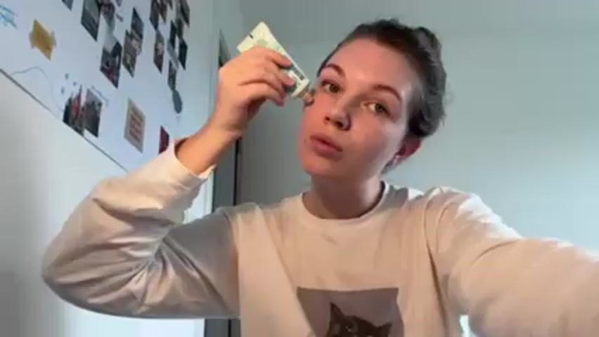 A woman with her hair in a bun is squeezing a tube of what appears to be cream or foundation onto her face, specifically near her eyebrow and cheek. She is looking directly at the camera. She is wearing a long-sleeved white shirt with a graphic image of a black cat on it. Behind her, a wall is decorated with photos and notes. The lighting appears to be natural daylight.