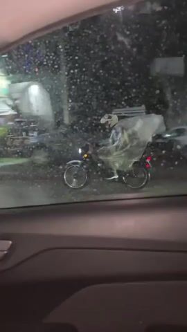 From within a vehicle, a person wearing a white helmet and a large, clear plastic covering rides a small motorbike along a wet, rain-slicked street. Street lights and building lights are visible in the background, blurred by the rain. The vehicle from which the video is shot appears to be moving.