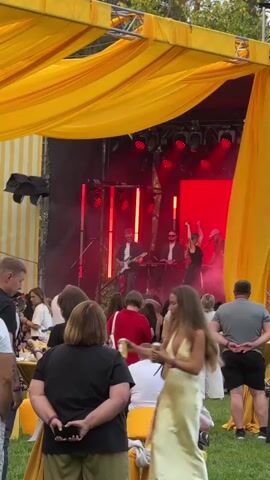 A band performs on a stage draped with yellow fabric. The stage is lit with red lights. The band consists of a guitarist on the left, a keyboardist in the center, a female singer in the center-right, and another musician on the right, possibly playing guitar. The singer is wearing a black top and white pants and is dancing. There is a crowd of people watching the performance. The environment appears to be outdoors.