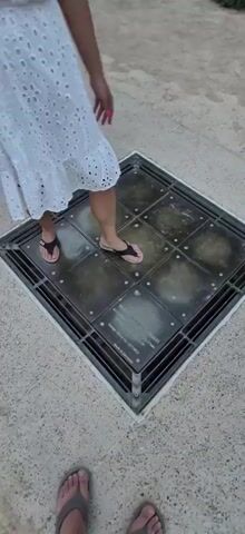 A person wearing a white dress and flip-flops is standing on a square grate with glass panels. The person is in the process of stepping onto the grate. The grate is embedded in a concrete surface. The person's right foot is on the grate, and their left foot is about to step down. The person is holding a pink object in their right hand. Another person's bare feet are visible in the bottom left corner of the frame. The video appears to be taken outdoors during the day.