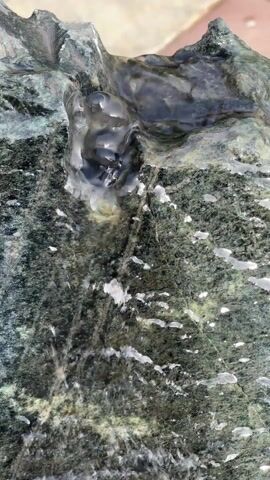The video shows water flowing over a rock feature. The rock is dark, with a coarse, speckled texture and lighter-colored veins. The water flows from a small pool at the top, through a narrow channel, and cascades down the rock surface. Small droplets of water are scattered across the rock. The background is slightly blurred, but appears to be a concrete surface and some greenery. The camera is close-up and stationary.
