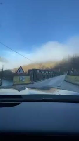 A car is driving onto a steel bridge, the road appears to be covered in ice or frost. There is fog in the distance and sparse trees. The sky is blue.