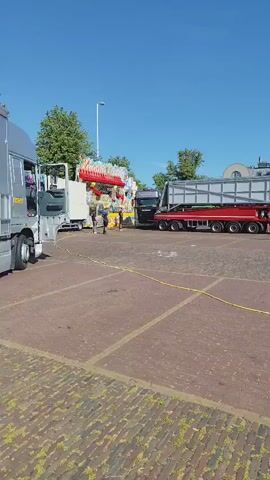 The video pans across an outdoor paved area under a bright blue sky with scattered clouds. Several large trucks, including semi-trailer trucks, are parked in the area. One truck has a long flatbed trailer carrying a large, boxy, metal structure. Another truck appears to be a flatbed with a fairground ride, possibly a roller coaster component, loaded onto it, and another truck is a semi-trailer with the words "BREAK DANCE" emblazoned on its side in large, red and yellow letters. Two men, wearing casual clothing, are visible near one of the trucks, seemingly engaged in some activity related to setting up or moving the ride. A third person, also casually dressed, is seen taking a photo with a mobile phone. The area appears to be a public space, possibly a town square or parking lot, with buildings visible in the background. A yellow hose or cable runs across the paved surface in the foreground.
