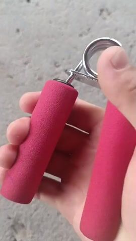 A person's hand is shown gripping a red hand exerciser. The hand is squeezing the exerciser repeatedly. The background appears to be a concrete surface.