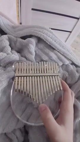 A hand holds a crystal kalimba, or thumb piano, on top of a gray blanket. The instrument has metal tines of differing lengths. The hand plucks the tines, producing sounds. A door with white panels is in the background.