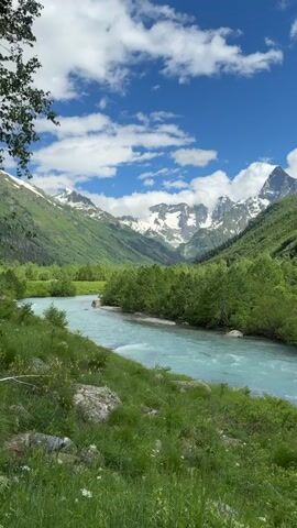 A serene mountain landscape unfolds with a clear blue sky dotted with white clouds. A turquoise river flows through a valley, flanked by lush green trees and rolling hills. In the distance, snow-capped mountains rise majestically against the horizon. The foreground features a grassy embankment with large rocks and scattered yellow wildflowers. The overall impression is one of natural beauty and tranquility.
