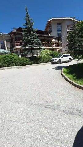 A white SUV is driving on a paved road. There is a building with balconies and trees in the background. The sky is clear and blue. The video is shaky, as if it was recorded by someone walking or running.