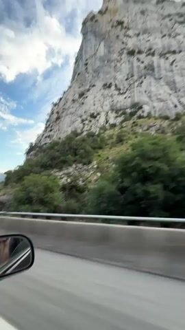 The video shows a POV shot from a car driving on a highway. The camera is looking out the side window towards the right. A large, rocky mountain covered in green foliage dominates the right side of the frame, with a blue sky and scattered clouds above. In the foreground, the side mirror reflects a person wearing a leopard-print dress, sitting in the passenger seat. The highway has multiple lanes, with a concrete barrier separating the lanes. Further in the distance, more mountains and trees are visible, along with power lines. The video has a slight motion blur, indicating the car is in motion.