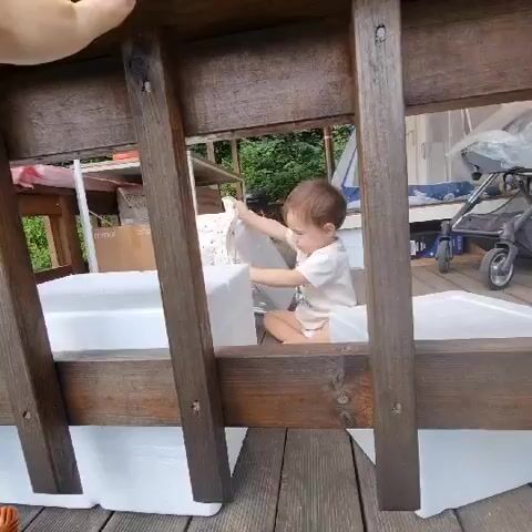 A baby is playing with white objects, possibly styrofoam boxes, inside a wooden structure. The baby is wearing a white onesie and is on their hands and knees. The structure appears to be outdoors on a wooden deck. In the background, there is a stroller, boxes, and some greenery. The camera is positioned to look through the wooden slats of the structure.