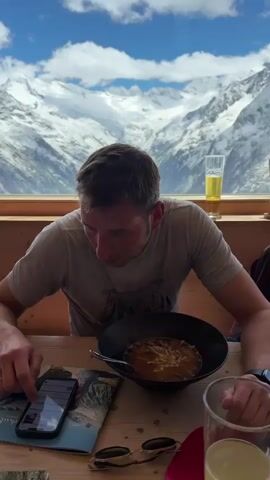 The video shows a man sitting at a wooden table, looking down at his phone. He is wearing a grey t-shirt and a watch on his left wrist. In front of him is a bowl of soup with cheese on top, a spoon, a cell phone, a pair of sunglasses, and a glass of beer. In the background, through a large window, is a breathtaking view of snow-covered mountains and a lake. The man is interacting with his phone by touching the screen with his index finger. Another glass, partially visible on the right side of the frame, appears to contain a red liquid. The overall scene depicts a person enjoying a meal and a drink with a stunning natural landscape as a backdrop.