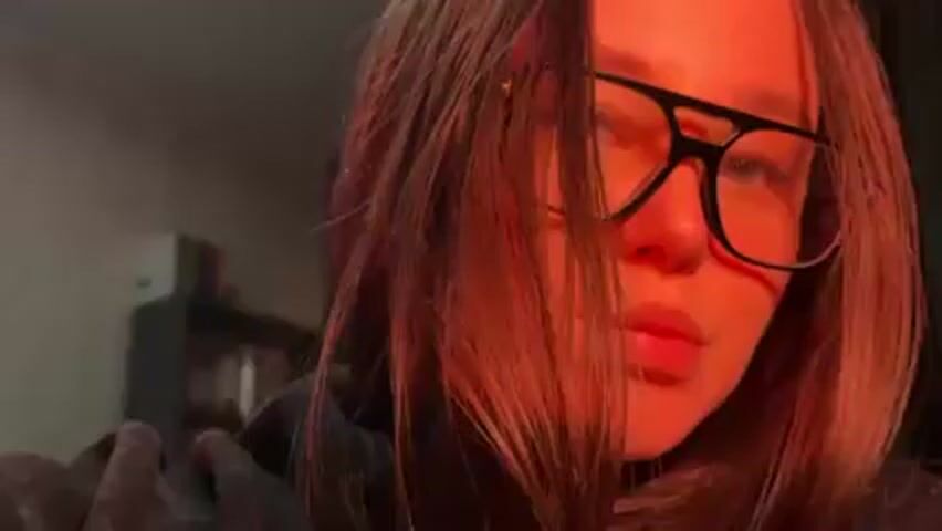 A person with brown hair and glasses is indoors with red lighting. The person is wearing a gray hoodie and looking slightly to the side. A blurred shelf is visible in the background.