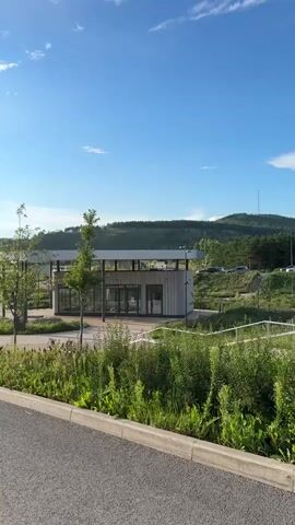 The video shows a paved pathway winding through a green landscape with a body of water to the left and rolling hills in the distance under a bright blue sky with scattered clouds. A modern-looking building with glass doors is visible on the left side of the path. Several young trees are planted along the pathway, and the ground is covered with lush green grass and wild plants. In the middle of the video, a person is seen riding a bicycle away from the camera on the pathway, casting a long shadow. The sun is shining brightly, creating a warm, summery atmosphere.