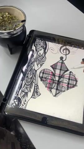 Here is a detailed description of the video:

The scene shows a person using a digital pen on a tablet. On the tablet screen is a digital drawing of what appears to be a hindu deity, along with playing card suits shaped like a heart, diamond, and club. The card suits are patterned with a plaid design. A small metallic pot containing a beverage with green herbs is in the upper left corner of the frame, to the left of the tablet. The surface the tablet sits on is white.