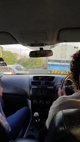 The interior of a car is seen from the back seats. A person with curly hair is driving the car. A person in the passenger seat is holding a cell phone. The car is at a standstill, waiting behind another car at an intersection. There is a large truck with a ladder on the back directly in front of the car.