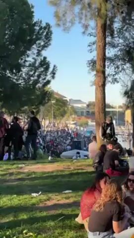 The video depicts a large group of people gathered in a park-like setting on a sunny day. Many individuals are sitting or lying on the grass, while others are standing and interacting. Some people are in small groups, talking and socializing. A large crowd is visible in the distance, suggesting a possible event or gathering taking place. Trees dot the landscape, providing shade to some of the park's occupants. A body of water is visible in the background, adding to the scenic nature of the location. The overall atmosphere appears relaxed and social, with people enjoying the pleasant weather and each other's company.
