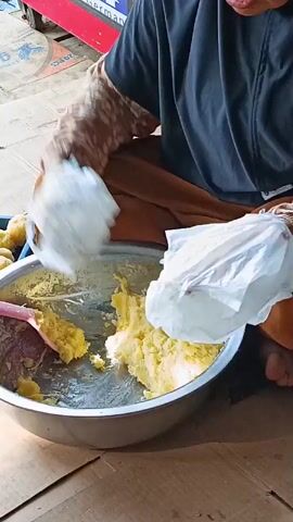 A person wearing a hijab and a patterned long-sleeved shirt is sitting and mashing yellow food in a large metal bowl with a pink spatula. The person is also holding two white plastic bags over the bowl, appearing to be adding something from the bags to the bowl. To the left of the bowl, there is a blue plastic container filled with round, yellow food items, possibly potatoes. The scene appears to be outdoors, with a red and white cart in the background. The ground is covered with cardboard.