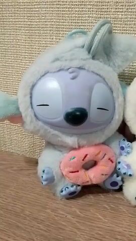The video shows two Stitch plush toys dressed in fluffy hooded outfits, sitting next to each other on a wooden surface. The Stitch on the left is wearing a light blue outfit and holding a pink donut. Its eyes are closed. The Stitch on the right is wearing a white outfit and holding a yellow pineapple. Its eyes are open and have star-shaped reflections inside them. The background is a textured beige surface. The camera focuses on the toys and provides a close-up view of their details. The scene is brightly lit.