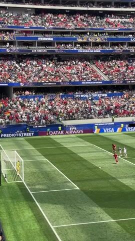 An aerial view of a soccer match in a stadium. A player in a red uniform is about to kick a penalty shot. Several other players in red uniforms are in the penalty box. One player in a white uniform is the goalkeeper. Spectators fill the stands, dressed in red and white. Banners with logos such as FIFA WORLD CUP 2026, QATAR AIRWAYS, and VISA are visible.