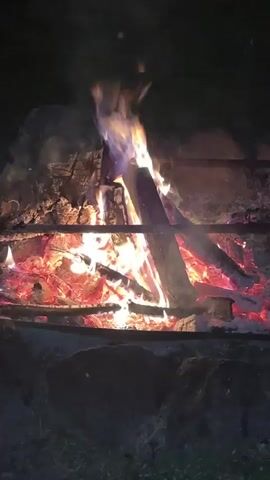 A close-up video shows a vibrant campfire burning in the dark. Large logs are consumed by bright orange and yellow flames, with hints of blue visible at the base of the fire. Beneath the active flames, a bed of red-hot embers glows intensely. Smoke gently rises from the fire, and small sparks can be seen drifting upwards. Two dark metal bars run horizontally across the top of the fire, suggesting a fire pit or a simple grill setup. The surrounding area is dark, indicating it is nighttime.