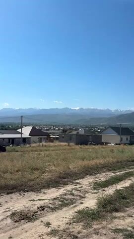 A vertical, wide shot captures a rural landscape under a clear blue sky. In the foreground, a dirt path cuts through dry, overgrown grass. The middle ground features several houses, some partially constructed and others complete with roofs. The background is dominated by a range of majestic mountains with snow-capped peaks, receding into the distance. The overall impression is one of a serene, undeveloped area with a backdrop of impressive natural beauty.