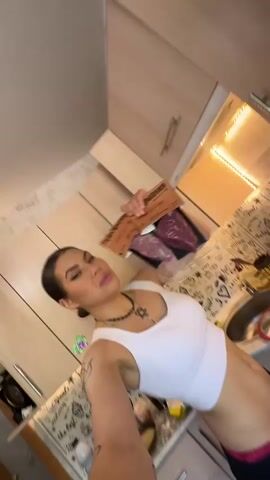 A medium shot captures a woman, wearing a white sports bra and dark pants, holding up a clear tray of packaged raw meat in front of a kitchen cabinet. She looks towards the camera with a slight smile. The camera then cuts to a close-up shot of her face as she speaks, then to a shot of two small, plastic-wrapped packages of meat and two larger brown packages, all with black cow silhouettes, resting on a white counter next to a white gas stove.