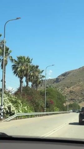 The video shows a car driving down a highway on a sunny day.  There are palm trees and other vegetation on the side of the road. A large hill or mountain is to the right of the road. There are other cars on the road. The video is taken from the inside of a car, looking out the windshield.
