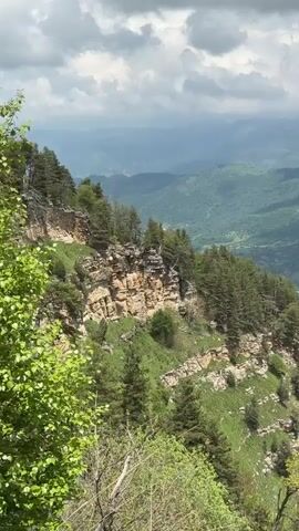 The video begins with a view of a steep, rocky cliff face covered in green foliage and trees. The camera pans to the right, revealing a vast valley with a winding river, a road, and a small village nestled among green hills. The landscape is lush and verdant, with rolling mountains stretching into the distance under a cloudy sky. The camera then shifts to focus on the branches of a large tree with green leaves.
